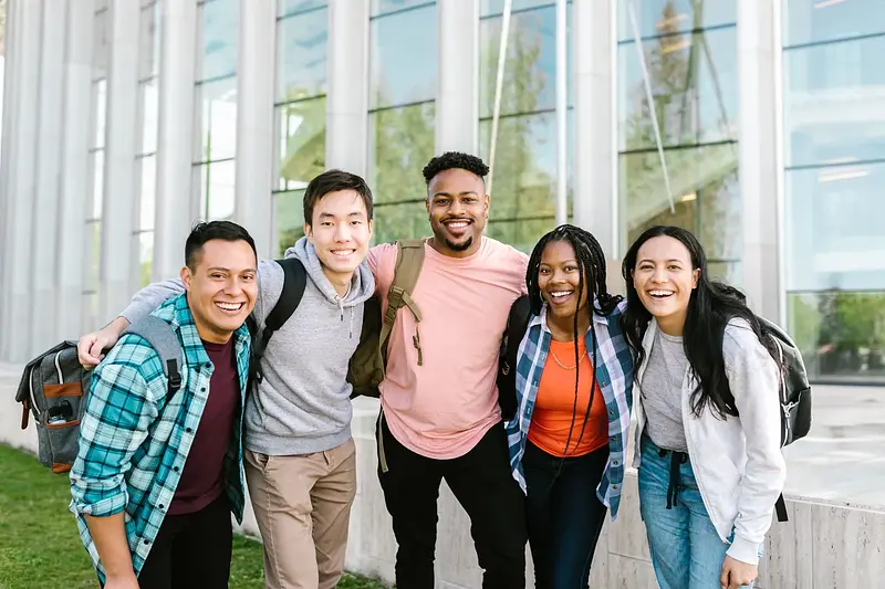Five smiling college students with backpacks standing arm in arm outside a campus building